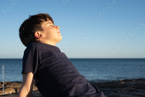 Young kid enjoying good weather on the coast, shore, breathing deep fresh sea air with eyes closed during sunset