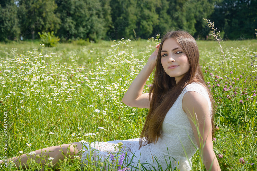 Beautiful young woman sits in a flowery meadow in a white dress