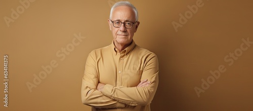 Waist up portrait of elderly man in glasses arms crossed looking at camera against beige background