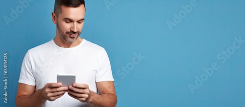A man in a white shirt uses an online app on his smartphone looking at the screen with a thoughtful expression standing isolated on a blue background © HN Works