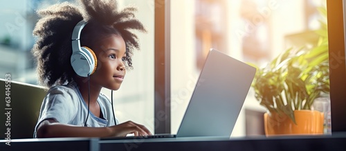 Young black girl with wireless headphones utilizing laptop at residence positioned by window engaged in activities involving computer empty area availa photo