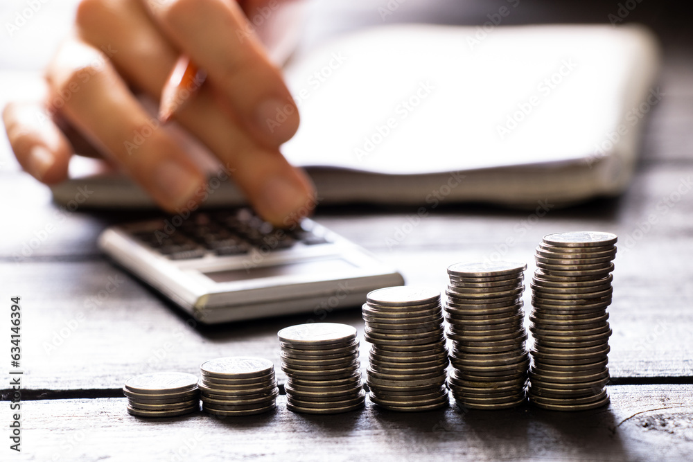 A hand on a calculator and next to it lies a notebook and stacks of coins, finance A hand on a calculator and next to it lies a notebook and stacks of coins, finance