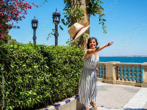 Caucasian tourist woman wearing a white and blue summer dress throwing her straw hat to the camera. Clara Campoamor Gardens with the Alameda Apodaca promenade in the background, Cadiz, Andalusia Spain photo