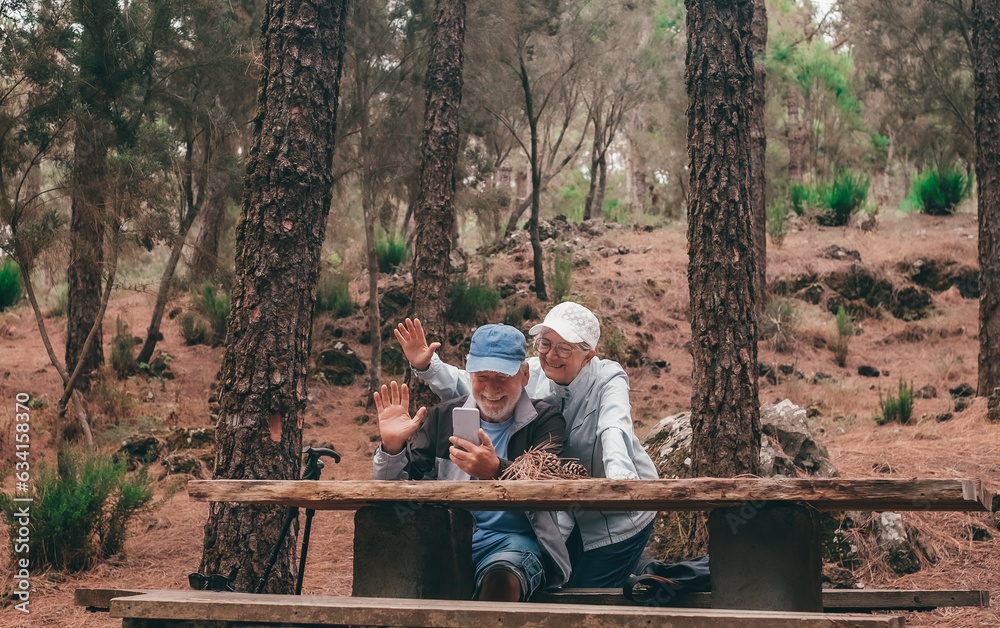 Video call concept. Happy senior couple in a trekking day in the woods using smatphone for online communication waving hands Video call concept. Happy senior couple in a trekking day in the woods using smatphone for online communication waving hands