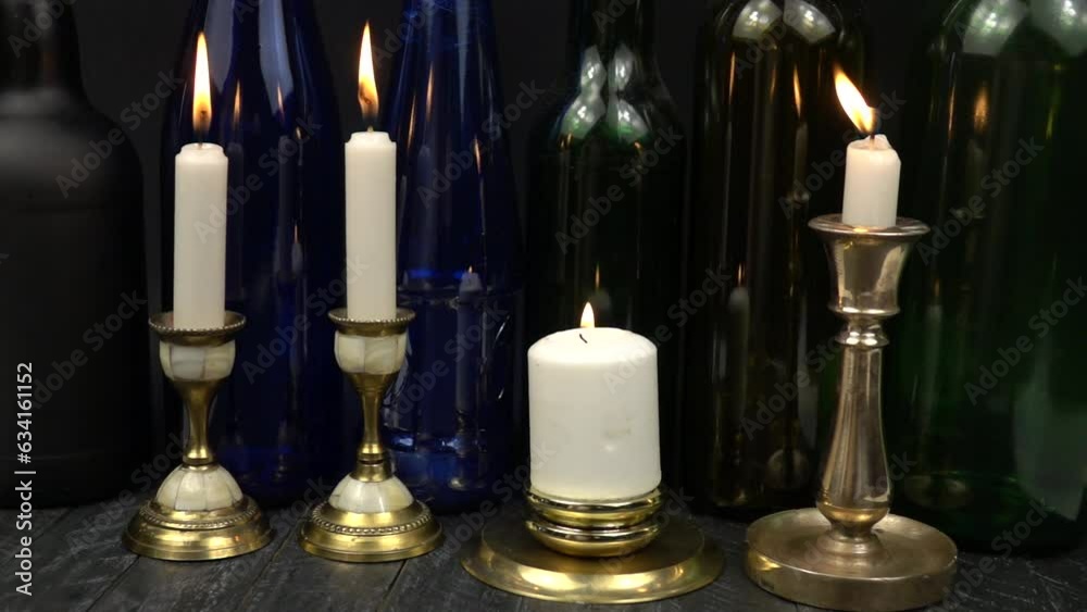 The flame of burning candles in two brass candlesticks is extinguished by a breath of wind. Burning white candles in old vintage brass and bronze candlesticks against the background of glass multicolo The flame of burning candles in two brass candlesticks is extinguished by a breath of wind. Burning white candles in old vintage brass and bronze candlesticks against the background of glass multicolo