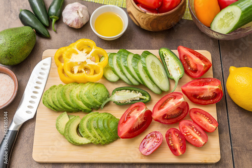 Different sliced vegetables for cooking salad on cutting board with knife on table on kitchen. Avocado, pepper, tomato, cucumber, oil, salt