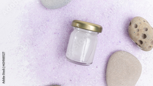 frasco de cristal transparente con tapa dorado con crema en fondo blanco con arena purpura y piedras decorativas para baño 