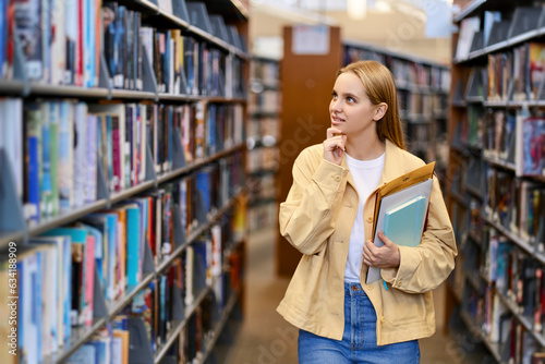Smart pretty focused blonde girl student holding books looking away walking in modern university campus library or bookstore thinking of college course study reading literature for doing research.