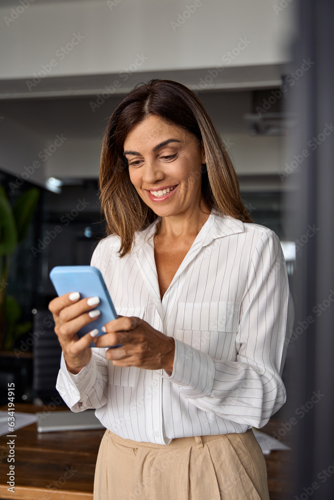 Vertical portrait of European businesswoman CEO holding smartphone using fintech application sitting on sofa indoors. Smiling Latin Hispanic mature adult professional business woman using mobile phone Vertical portrait of European businesswoman CEO holding smartphone using fintech application sitting on sofa indoors. Smiling Latin Hispanic mature adult professional business woman using mobile phone