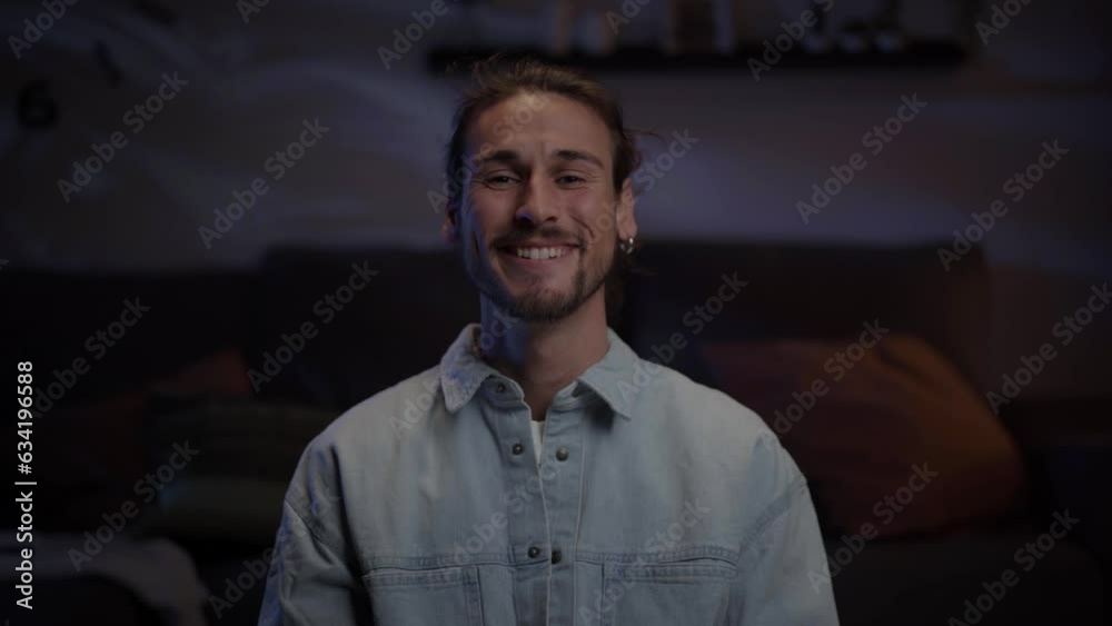 Attractive young Caucasian man looking smiling at camera indoor. European adult handsome man with beard poses happy in living room at night. Natural male beauty in millennial generation at home. Attractive young Caucasian man looking smiling at camera indoor. European adult handsome man with beard poses happy in living room at night. Natural male beauty in millennial generation at home.