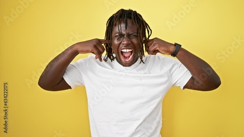 African american man screaming loudly covering ears with fingers over isolated yellow background photo