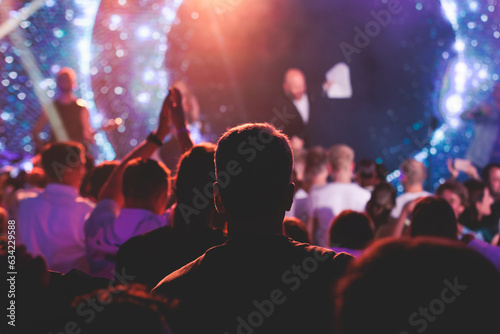 Crowded concert hall arena with scene stage lights with musicians band on a stage at the venue  rock show performance  with concert-goers attendees  audience on dance floor during concert festival