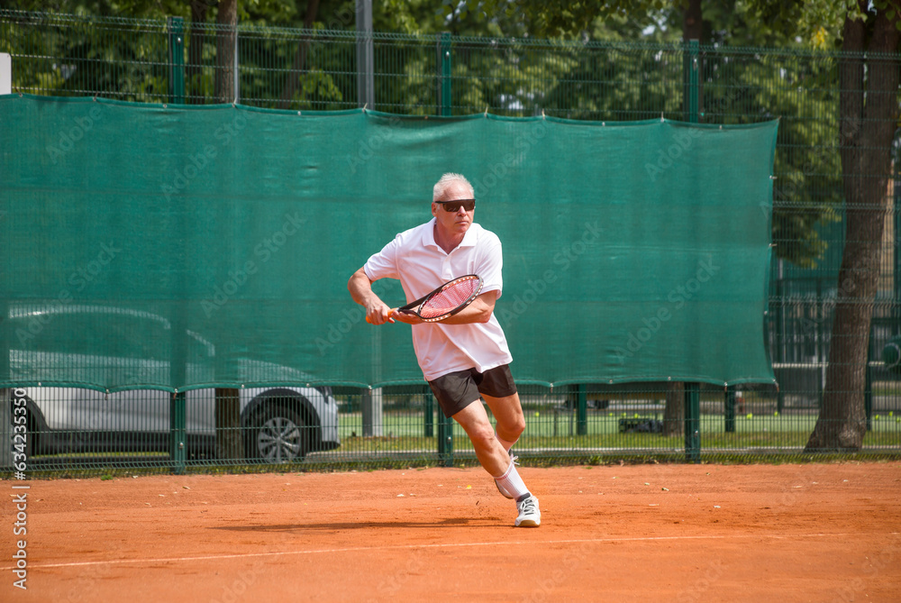 An elderly man playing tennis runs to take the ball. Open ground.