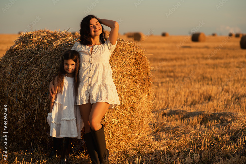  sweet little girl of European appearance hugs her mother tightly at sunset amidst a mowed field of wheat and hay bales