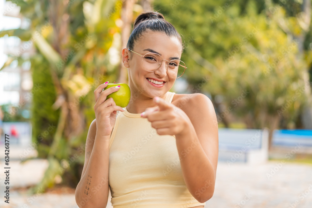 Young pretty woman with an apple at outdoors points finger at you with a confident expression Young pretty woman with an apple at outdoors points finger at you with a confident expression