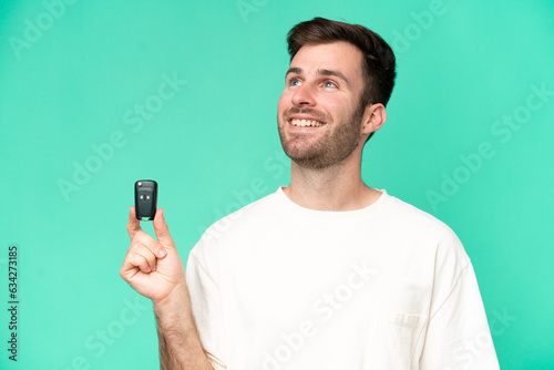 Young caucasian man holding car keys isolated on green background looking up while smiling