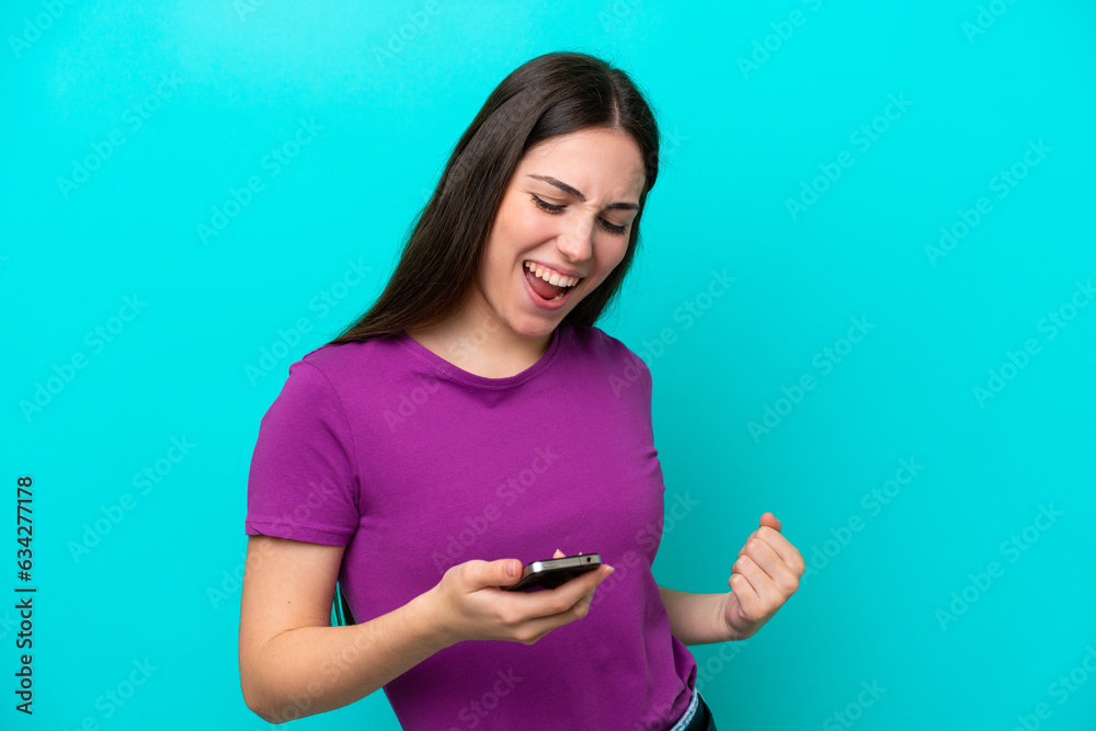 Young girl isolated on blue background using mobile phone and doing victory gesture Young girl isolated on blue background using mobile phone and doing victory gesture