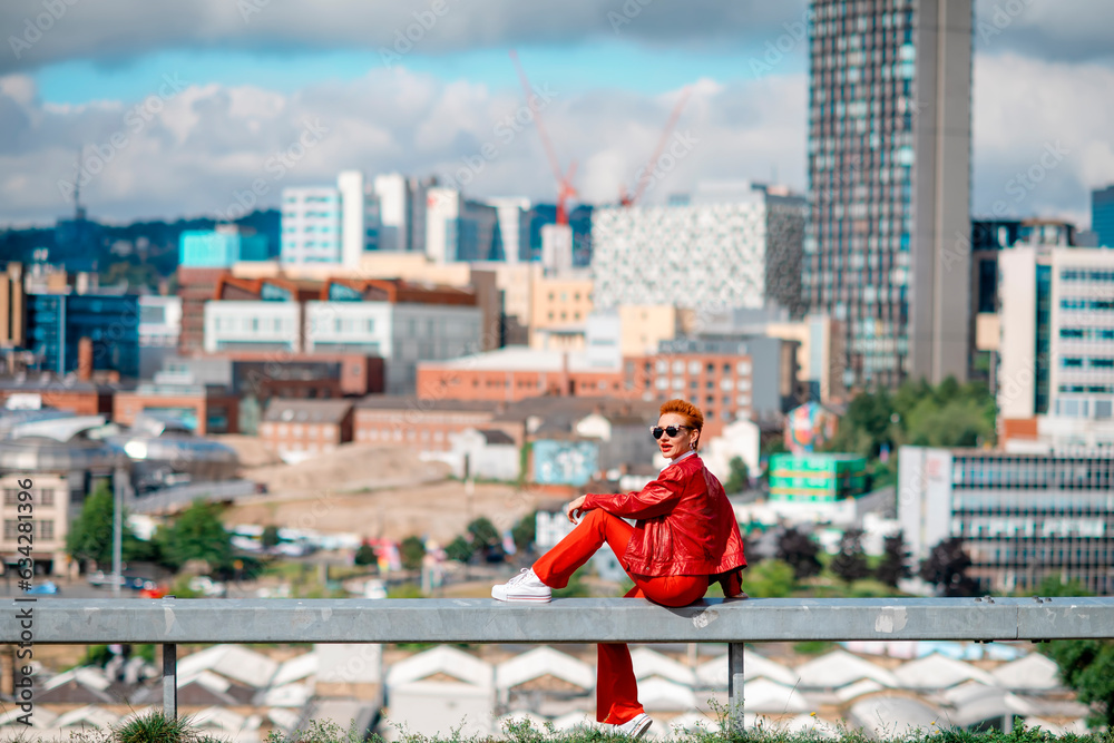 a red hair woman in a red costume sitting in front of the city on great summer day