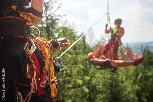 Paramedic with patient in stretcher under helicopter. Teams of emergency medical service and firefighters training rescue injured person from inaccessible terrain. Selective focus on safety harness.. photo