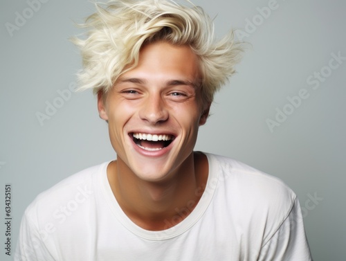 In this candid portrait, a man with golden locks stands in front of an indoor wall, joyfully laughing with a bright smile that reveals his pearly teeth and highlights his smooth skin and rosy cheeks