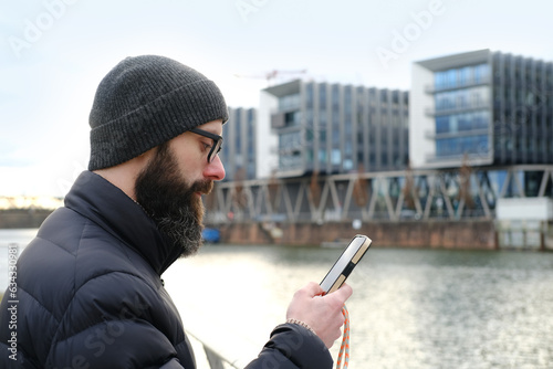 young bearded man in coat and hat stands on shore river Main, uses mobile phone, winter walks in Frankfurt, View of modern residential buildings, Gutleitviertel district on banks, concept of tourism photo