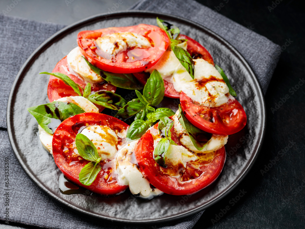 Plate of healthy classic caprese salad with ripe tomatoes and mozzarella cheese with fresh basil leaves on dark background. Close up