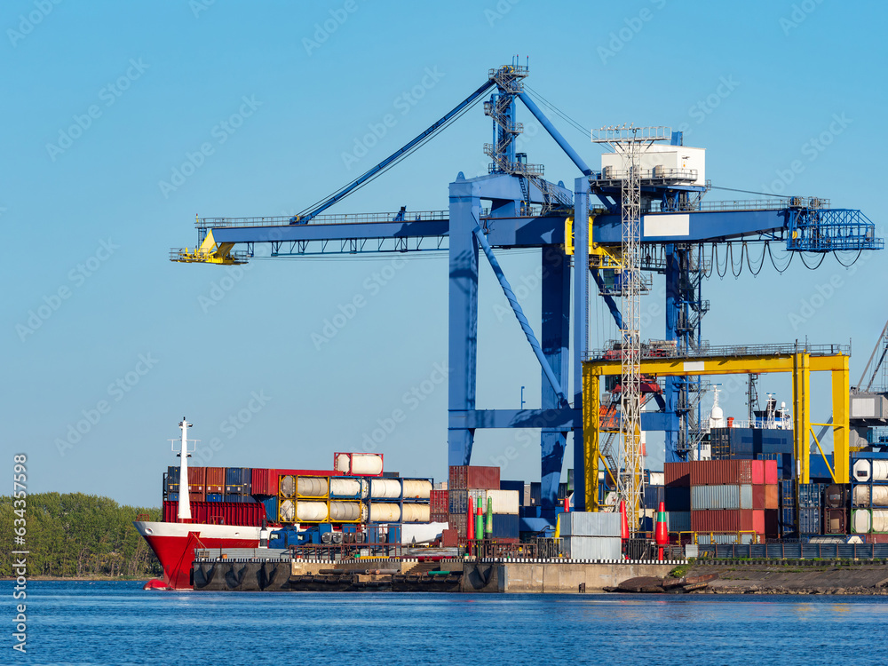 Marine logistics hub. Containers ship in cargo port. Sea barge is awaiting unloading. Cargo cranes load containers onto ship. Transportation of goods by sea. Port on summer day. Oceanic logistics