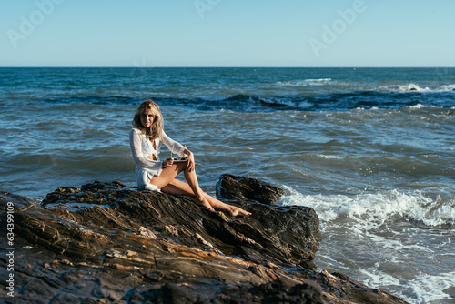 A beautiful girl in white glasses and white clothes sits on the rocks on the seashore