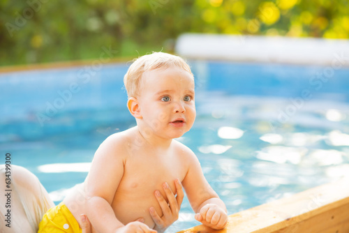 Portrait of small red-haired boy bathes in pool with hand support, baby swimming in water, summer leisure