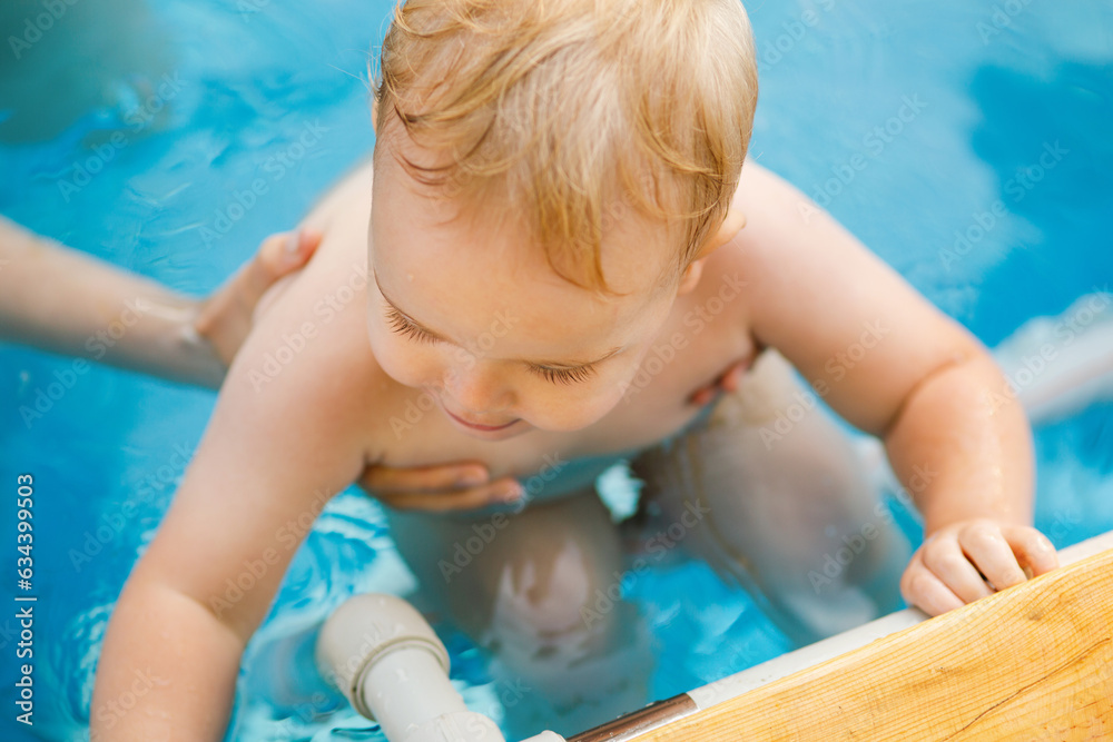 Portrait of small red-haired boy bathes in pool with hand support, baby swimming in water, summer leisure Portrait of small red-haired boy bathes in pool with hand support, baby swimming in water, summer leisure