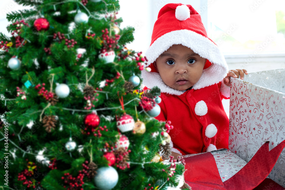 Cute baby African girl in red costume standing in big box near Christmas tree, looking at camera