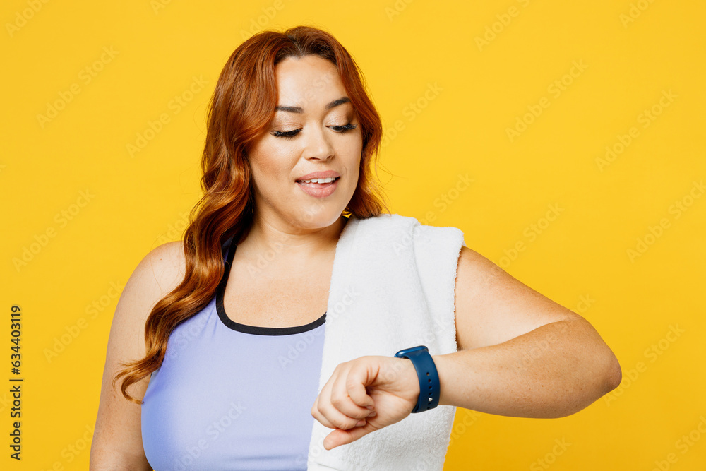 Young smiling satisfied chubby overweight plus size big fat fit woman wear blue top warm up training looking at smart watch isolated on plain yellow background studio home gym. Workout sport concept. Young smiling satisfied chubby overweight plus size big fat fit woman wear blue top warm up training looking at smart watch isolated on plain yellow background studio home gym. Workout sport concept.