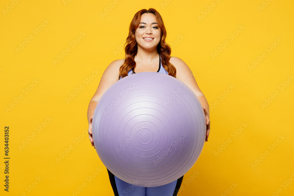 Young fun chubby overweight plus size big fat fit woman wear blue top warm up training hold in hand fit ball looking camera isolated on plain yellow background studio home gym. Workout sport concept. Young fun chubby overweight plus size big fat fit woman wear blue top warm up training hold in hand fit ball looking camera isolated on plain yellow background studio home gym. Workout sport concept.