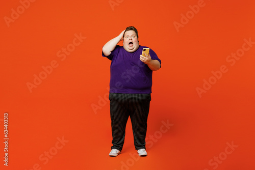 Full size body sad shocked young chubby overweight man wear purple t-shirt casual clothes hold in hand use mobile cell phone put hand on head isolated on plain red orange background Lifestyle concept