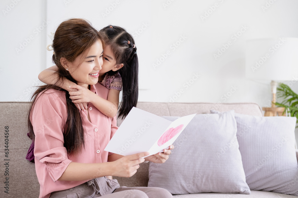 Cheerful mother hugging daughter and reading handmade greeting card with heart while resting on sofa couch during holiday celebration mothers day at home Cheerful mother hugging daughter and reading handmade greeting card with heart while resting on sofa couch during holiday celebration mothers day at home