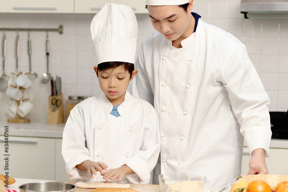 Happy Asian son and father in chef uniform with hat cook meal together at kitchen, dad parent and boy child prepare bread dough, cute family making bakery food. Kid student learns to bake a bread.