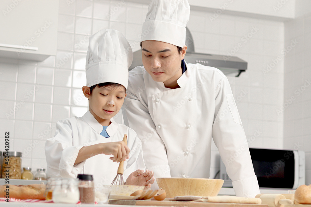 Happy Asian son and father in chef uniform with hat cook meal together at kitchen, dad parent and boy child mixing and kneading bread dough, cute family making bakery. Kid student learns to bake bread