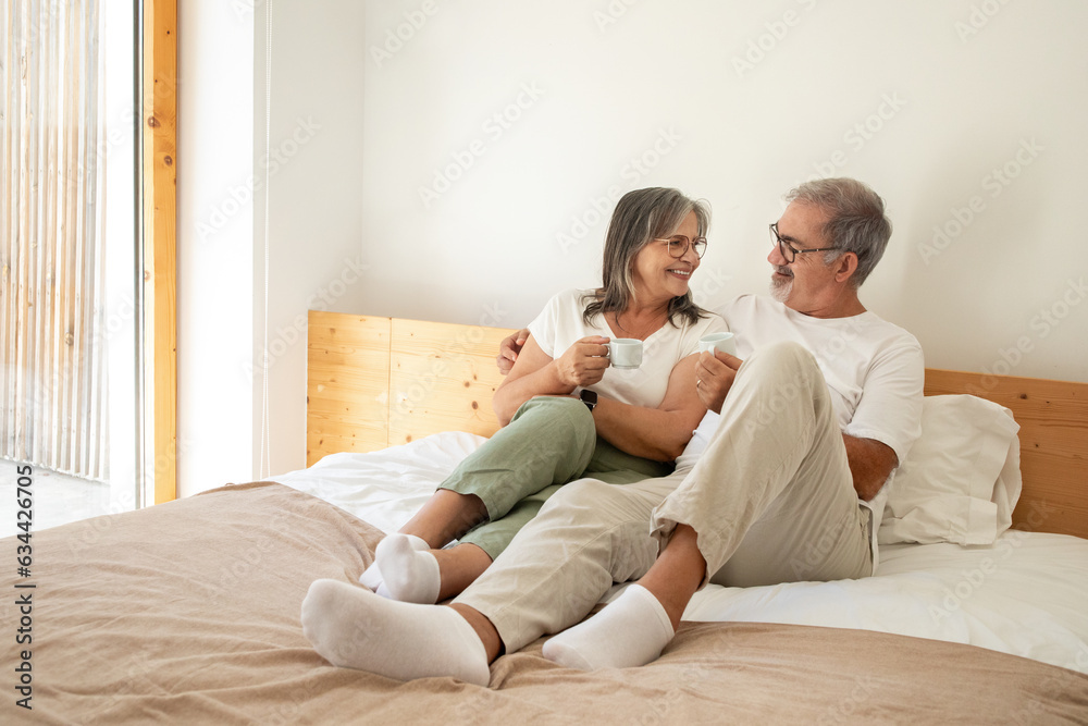 Cheerful old european man and lady have breakfast with cups of coffee, enjoy free time, sit on bed in bedroom interior