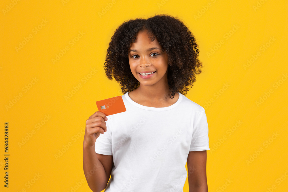 Cheerful adolescent black girl in white t-shirt, uses credit card, isolated on orange background Cheerful adolescent black girl in white t-shirt, uses credit card, isolated on orange background