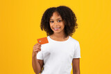 Cheerful adolescent black girl in white t-shirt, uses credit card, isolated on orange background