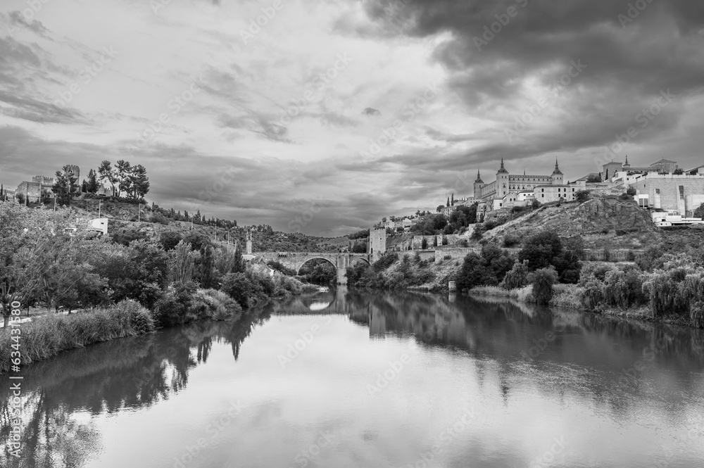 Black and white view of the Spanish city of Toledo, a UNESCO World Heritage Site, with the Tagus river and the Alcazar, under a threatening stormy sky