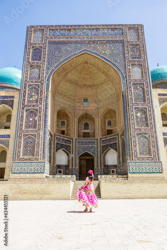 Young woman in traditional Uzbek dress at Bukhara, Uzbekistan Mir-i-Arab Madrasa Kalyan minaret and tower. Translation on mosque: "Poi Kalyan Mosque"