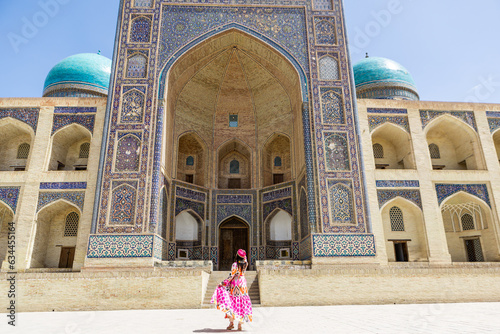 Young woman in traditional Uzbek dress at Bukhara, Uzbekistan Mir-i-Arab Madrasa Kalyan minaret and tower. Translation on mosque: "Poi Kalyan Mosque"