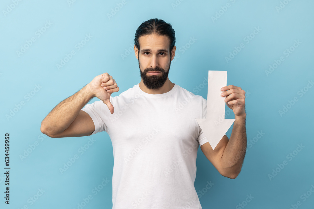 Portrait of disappointed sad handsome man with beard wearing white T-shirt showing white arrow pointing down, showing thumb down, dislike gesture. Indoor studio shot isolated on blue background. Portrait of disappointed sad handsome man with beard wearing white T-shirt showing white arrow pointing down, showing thumb down, dislike gesture. Indoor studio shot isolated on blue background.