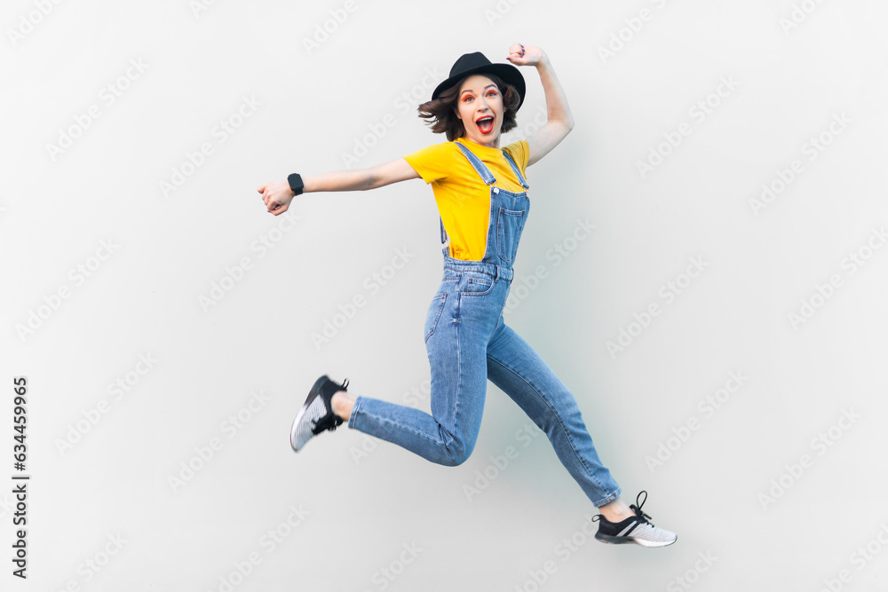 Portrait of excited positive hipster woman in blue denim overalls, yellow T-shirt and black hat, jumping in the air, moving, expressing happiness. Indoor studio shot isolated on gray background.