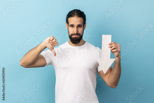 Portrait of disappointed sad handsome man with beard wearing white T-shirt showing white arrow pointing down, showing thumb down, dislike gesture. Indoor studio shot isolated on blue background.