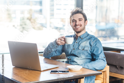 Portrait of delighted smiling happy young man freelancer in blue jeans shirt working on laptop, having break, drinking coffee. Indoor shot near big window, cafe background.