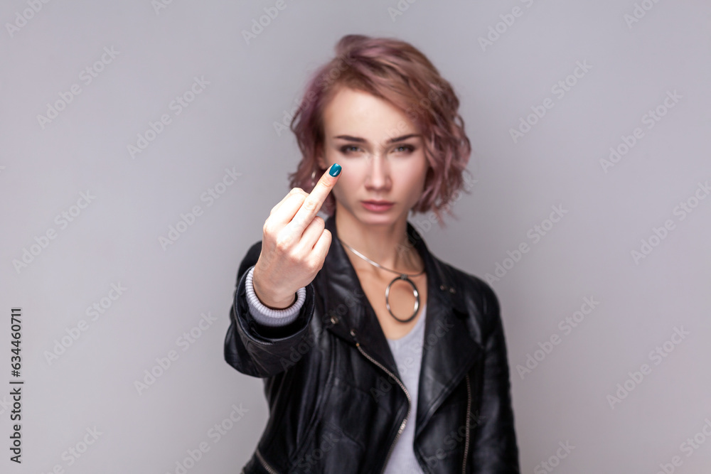 Portrait of serious rude woman with short hairstyle standing showing middle finger, impolite gesture, looking at camera, wearing black leather jacket. Indoor studio shot isolated on grey background. Portrait of serious rude woman with short hairstyle standing showing middle finger, impolite gesture, looking at camera, wearing black leather jacket. Indoor studio shot isolated on grey background.