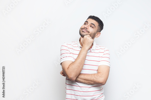 Portrait of pensive dreaming positive bearded man wearing striped t-shirt standing looking away, thinking about something pleasant. Indoor studio shot isolated on gray background.