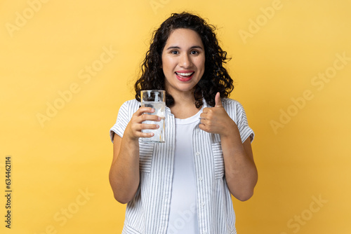 Portrait of smiling optimistic delighted beautiful woman with dark wavy hair standing with glass of water, showing thumb up, like beverage. Indoor studio shot isolated on yellow background.